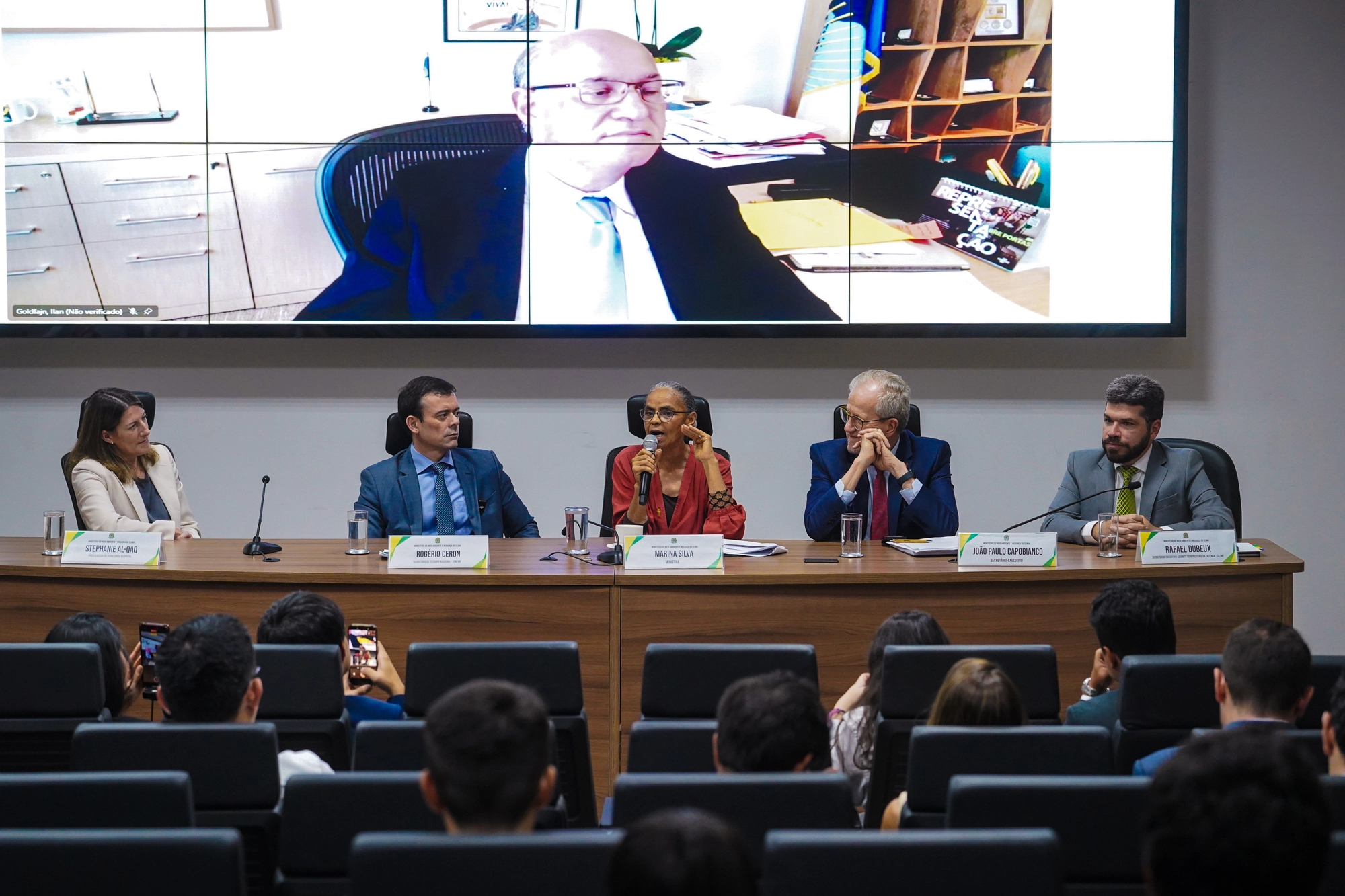 Painel de debate em um auditório com cinco participantes sentados atrás de uma mesa, enquanto uma pessoa participa por videoconferência em uma grande tela ao fundo. A mulher no centro fala ao microfone, e os demais participantes acompanham a discussão. Na plateia, pessoas assistem ao evento sentadas em poltronas, algumas registrando a cena com celulares. O ambiente é formal e institucional, com crachás de identificação sobre a mesa.