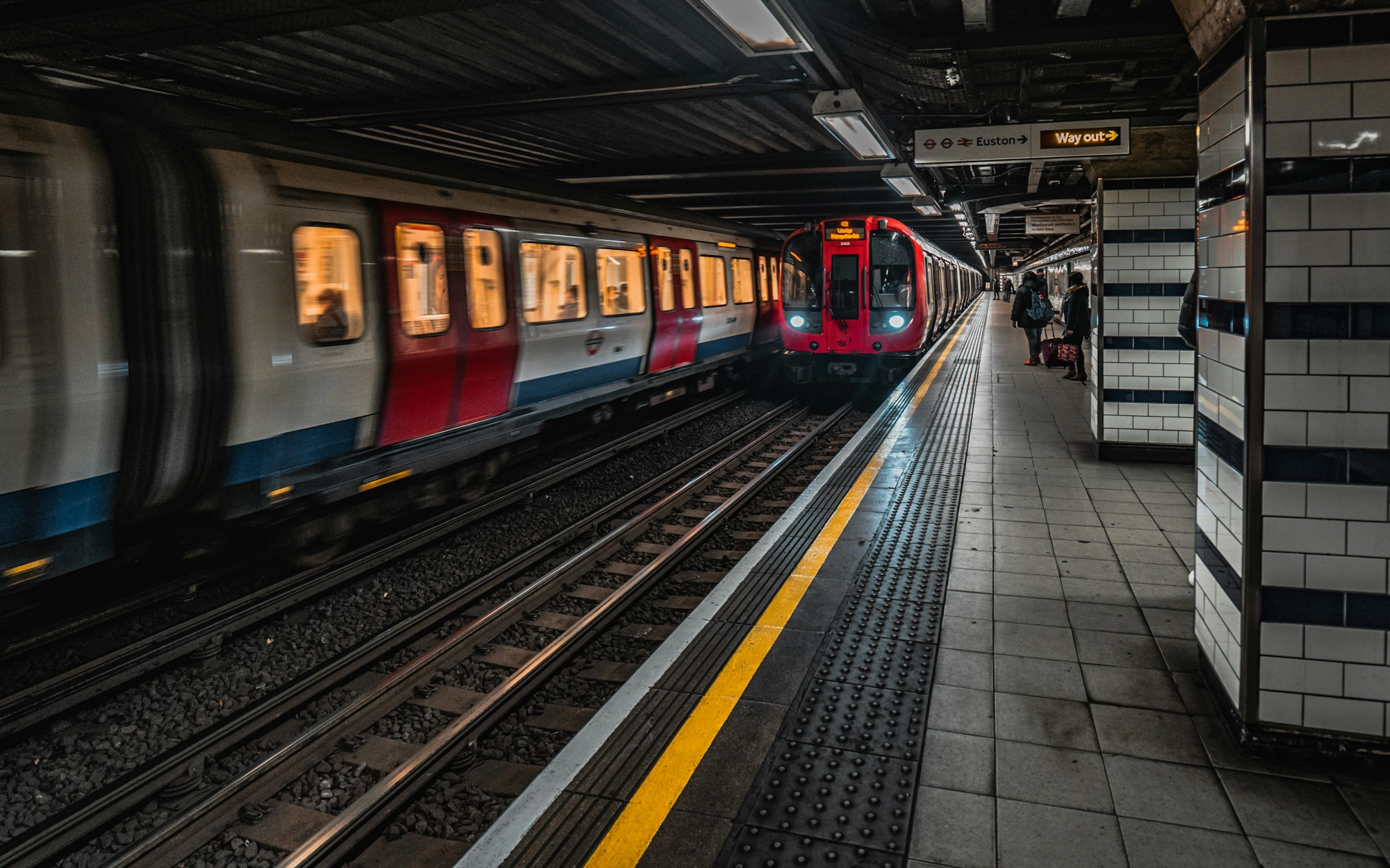 Estação de metrô subterrânea com dois trens em movimento sobre trilhos paralelos. Um trem vermelho se aproxima da plataforma, enquanto outro passa rapidamente no sentido oposto, criando efeito de desfoque. Algumas pessoas aguardam na plataforma iluminada, ao lado da faixa de segurança amarela, sob placas de sinalização suspensas e revestimentos de azulejos brancos e pretos nas paredes.